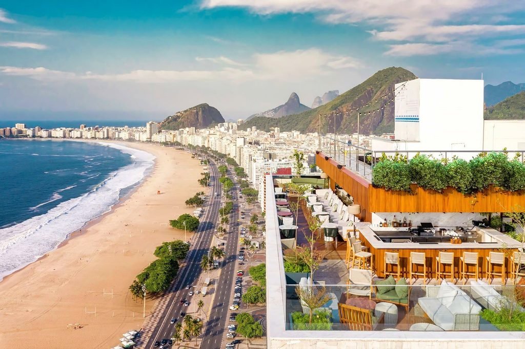 O foco é no Hotéis e na curva da Praia de Copacabana ao amanhecer, com a areia ainda vazia, o mar calmo refletindo tons de laranja e rosa, e o Pão de Açúcar imponente no horizonte. A imagem deve capturar a magnitude geográfica que justifica pagar por uma vista frontal no Rio de Janeiro